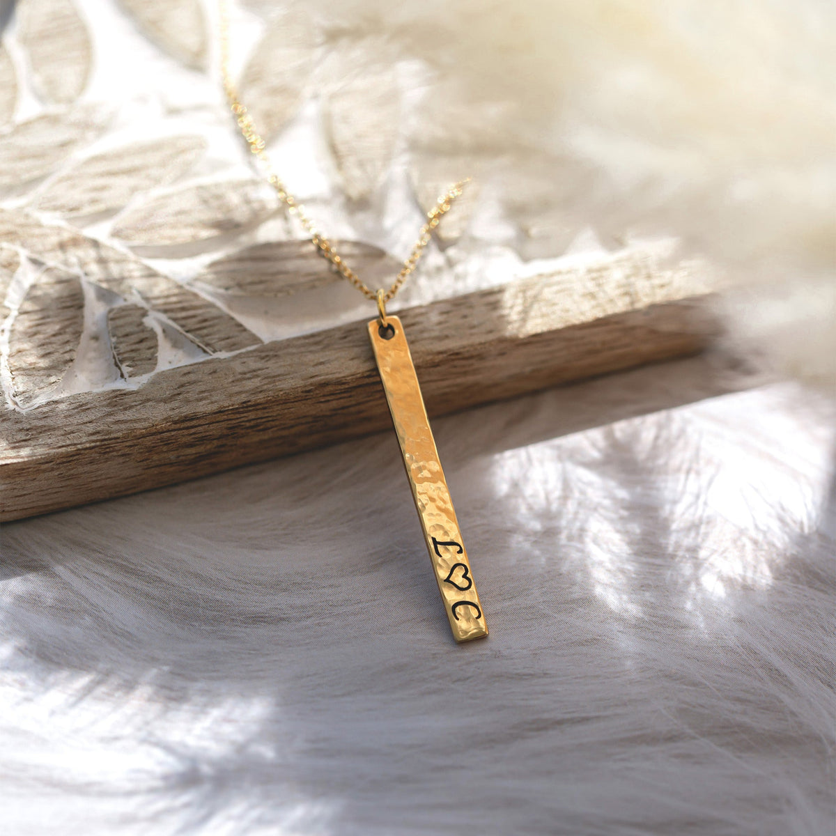 Gold-tone hammered vertical bar necklace engraved with initials and a heart symbol, displayed on white fabric