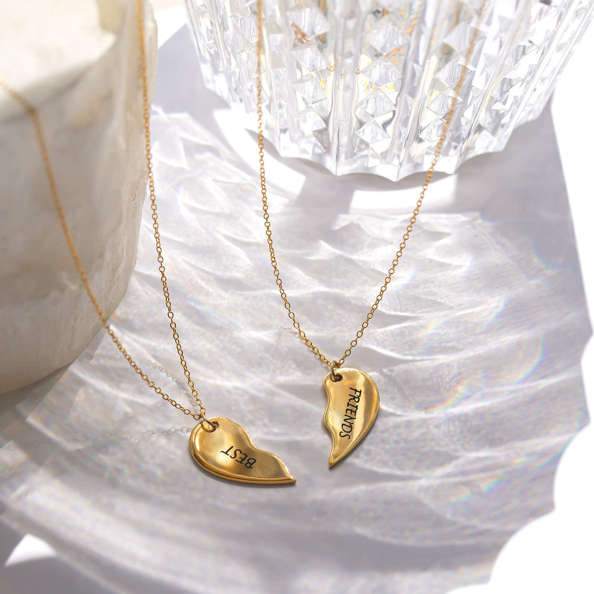 Gold-tone split heart necklaces engraved BEST and FRIENDS displayed on a reflective surface beside clear faceted decor