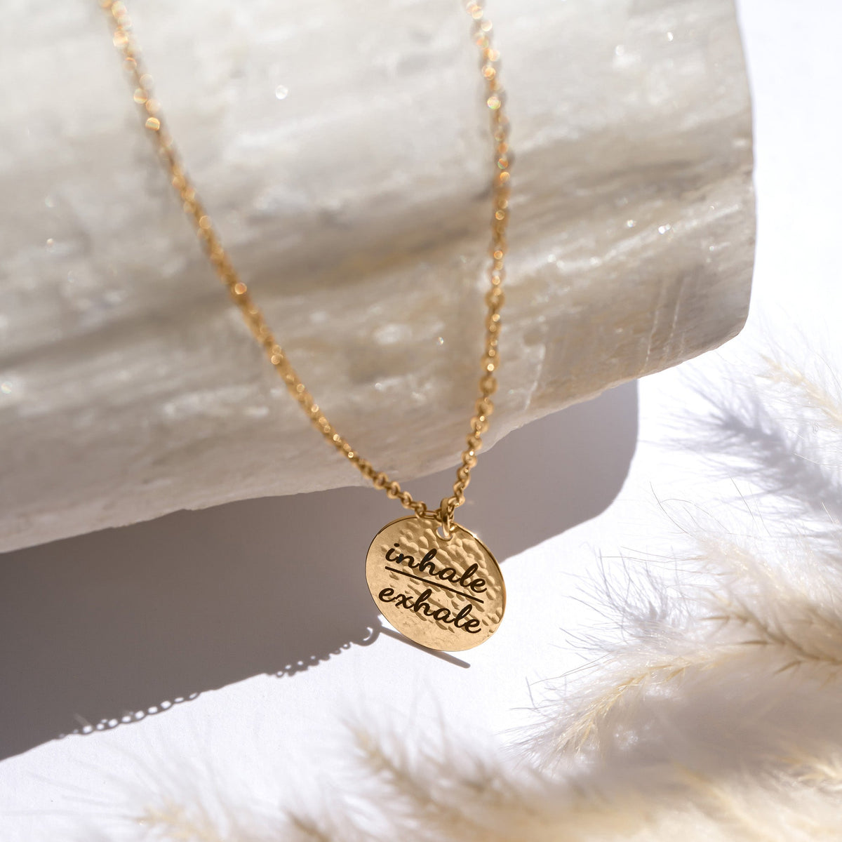 Gold-tone hammered round disc necklace engraved inhale exhale displayed beside a white textured surface