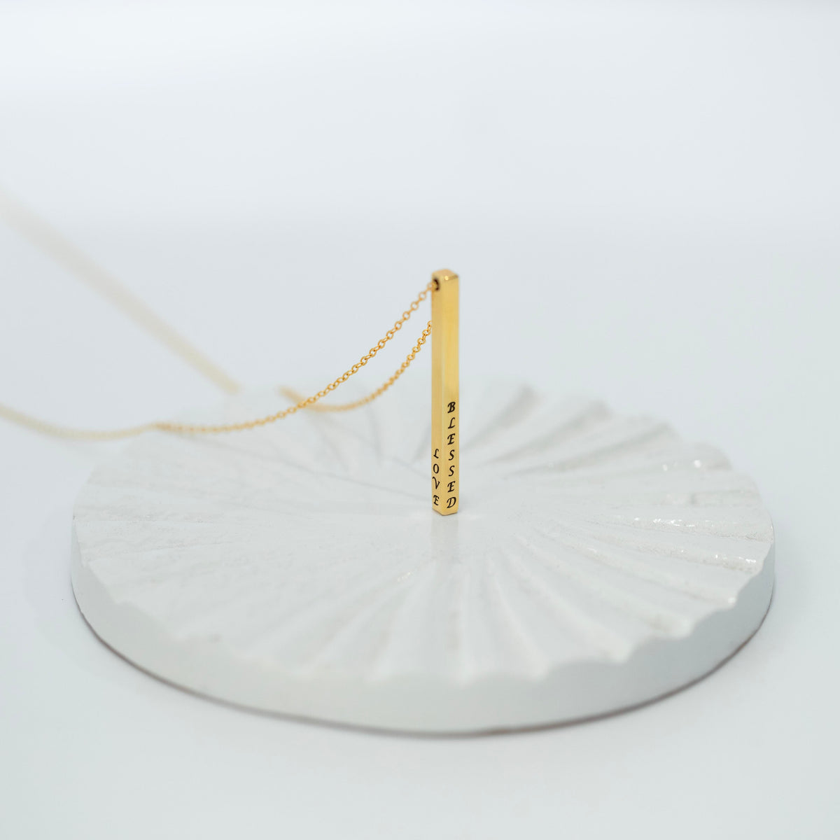 Gold-tone vertical bar necklace with engraved text on multiple sides, displayed on a white textured stand