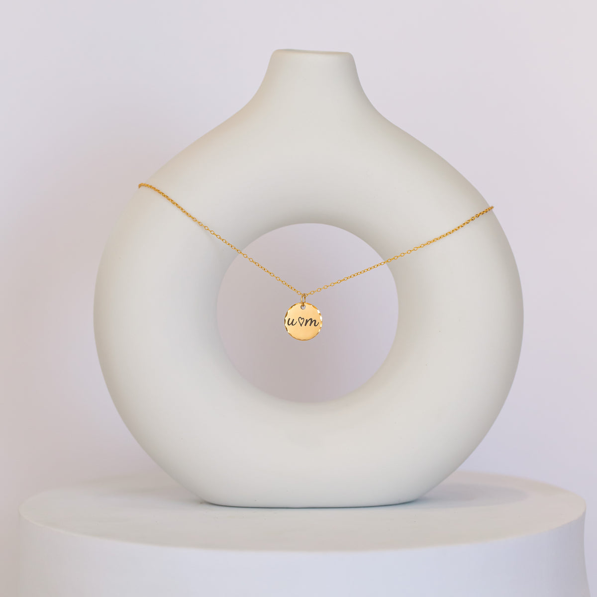 Gold-tone round disc necklace engraved U, a heart, and M displayed on a white sculptural stand
