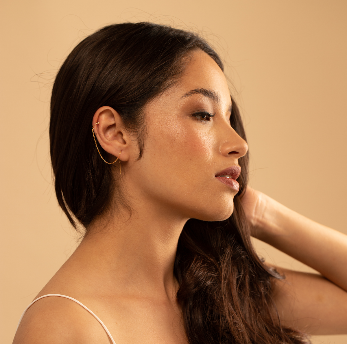 Woman with long dark hair and gold chain cuff earrings against a beige background