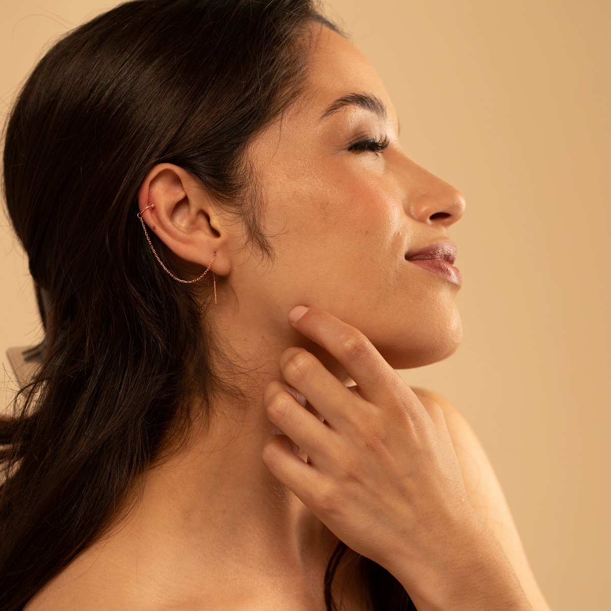 A woman with a gold threaded earring touching her neck with a beige background