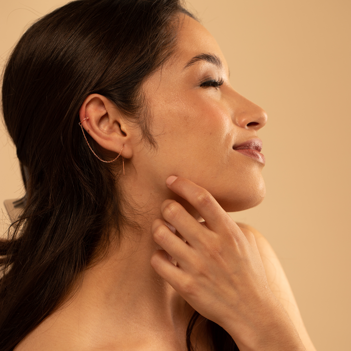 A woman with a gold threaded earring touching her neck with a beige background