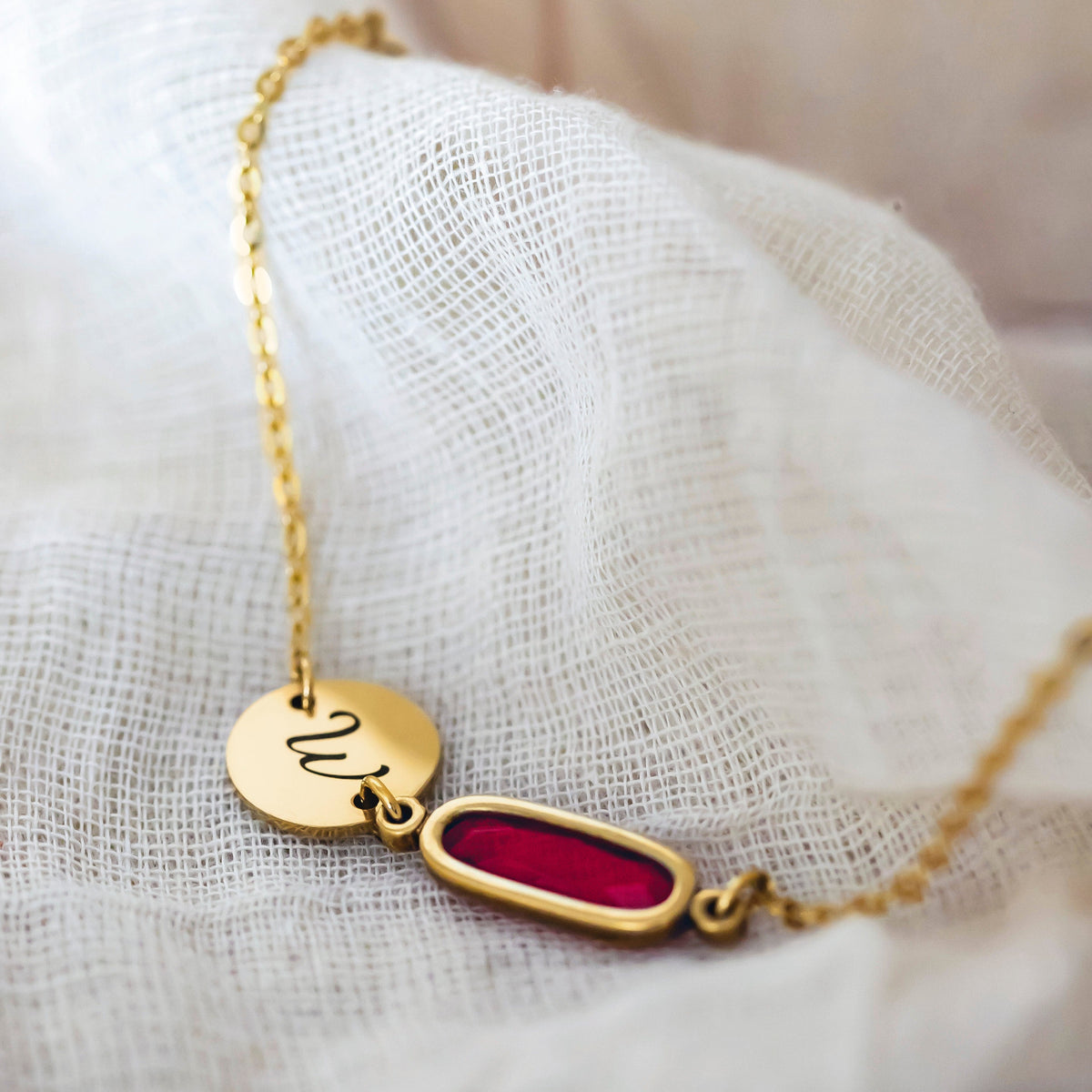 Gold-tone bracelet with engraved W disc and red elongated rectangular stone on white woven fabric