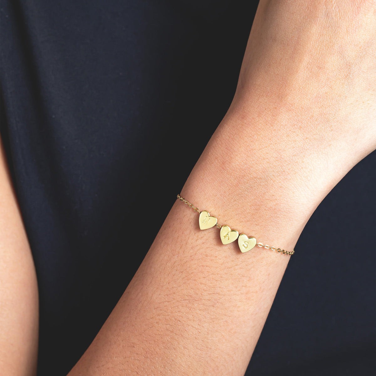 Gold-tone bracelet with three engraved heart charms worn on a wrist against a dark background