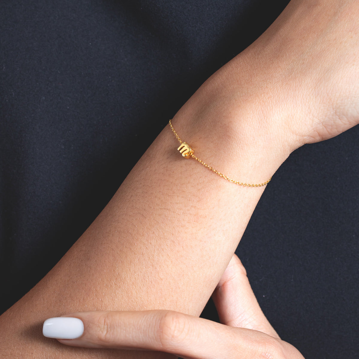 Delicate chain bracelet with small cursive initial charm worn on a wrist against a dark background