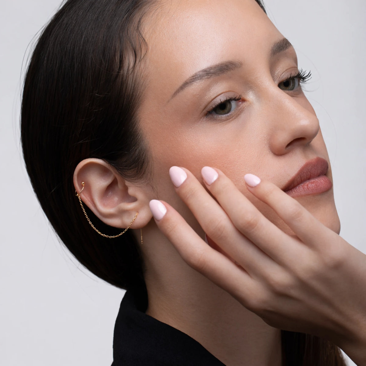 Close-up of a woman's face with hand near her face, wearing a gold chain cuff earring.