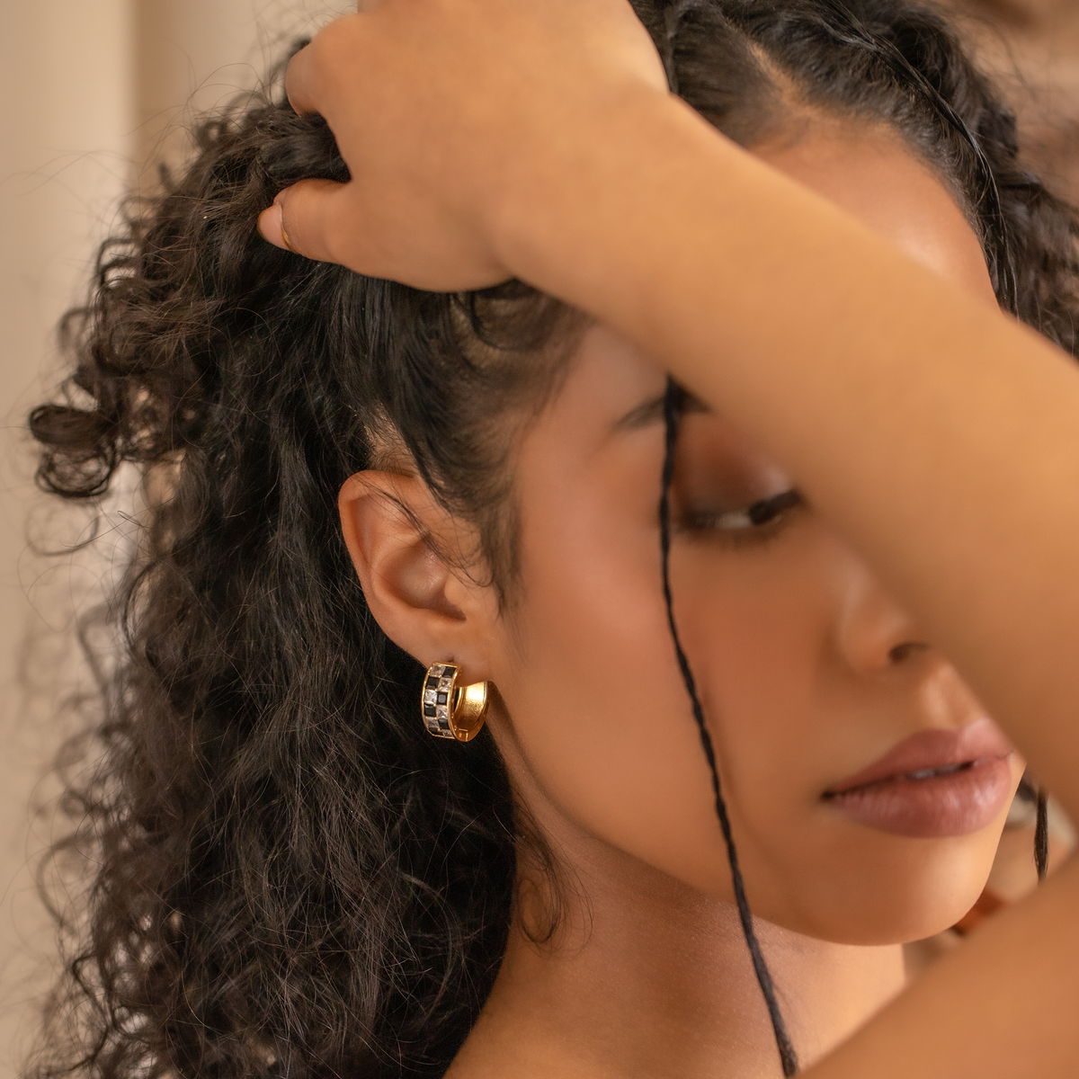 Close-up of a woman with her hand in her hair, wearing gold checkered design hoop earrings.