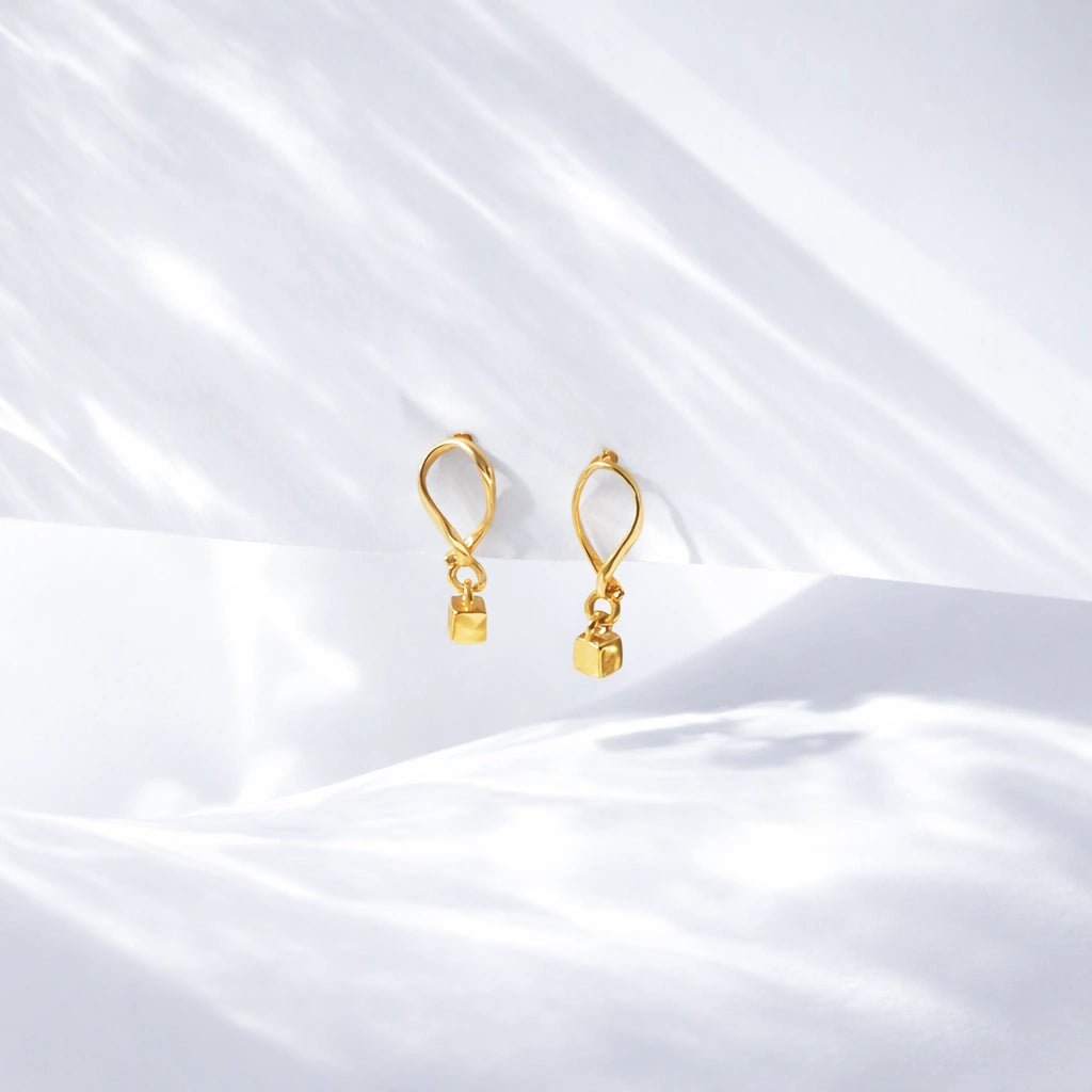 Gold-tone drop earrings with small square charms displayed on softly folded white satin