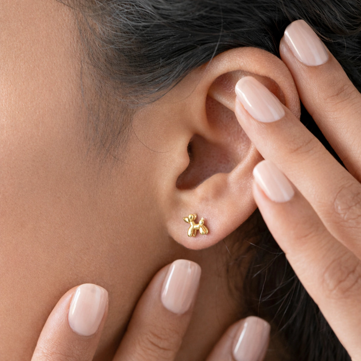 Close-up of a gold-tone balloon dog stud earring worn on the ear beside pale manicured nails