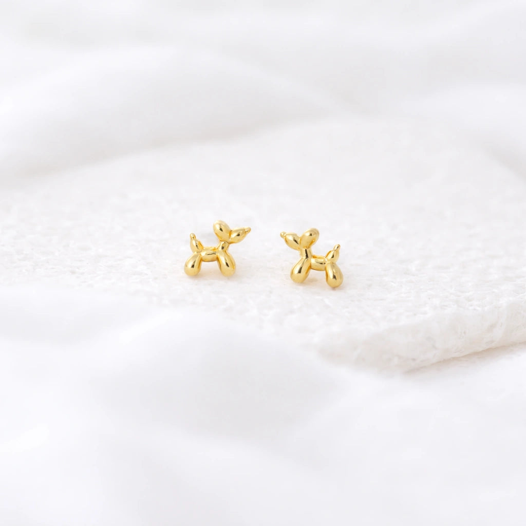 Gold-tone balloon dog stud earrings displayed on a soft white textured surface