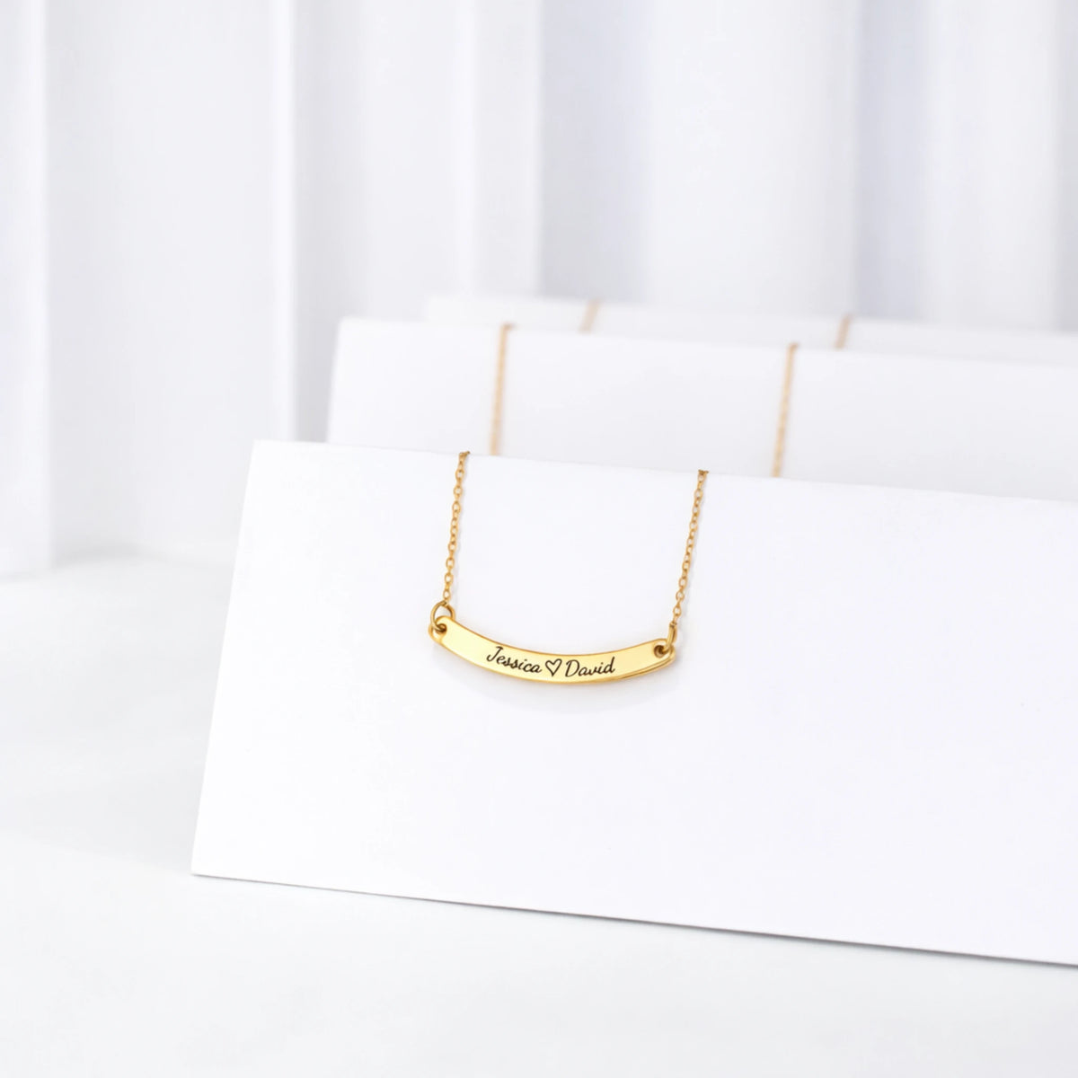 Gold-tone curved bar necklace engraved Jessica, a heart, and David displayed on white jewelry cards