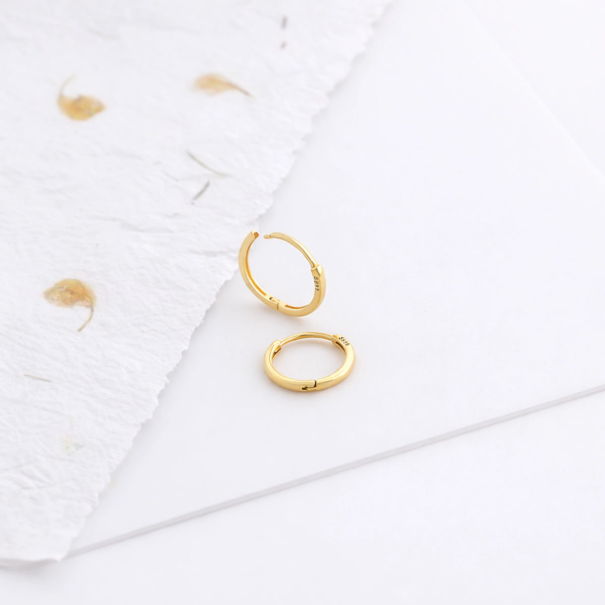 Gold-tone thin hoop earrings displayed on a white surface beside textured paper