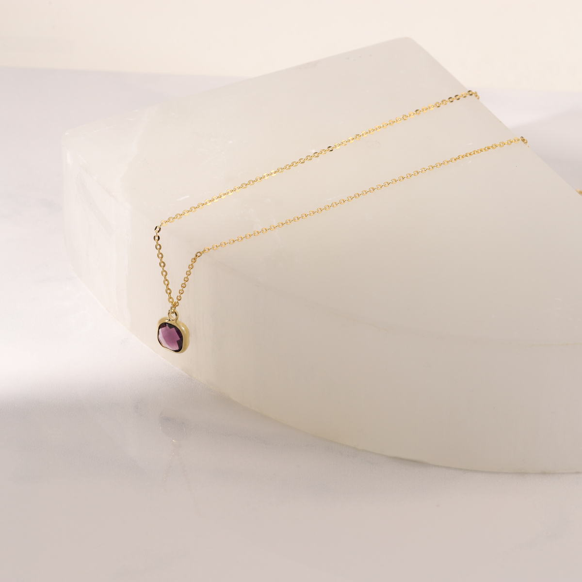 Gold-tone necklace with a purple faceted pendant displayed on a white curved stand