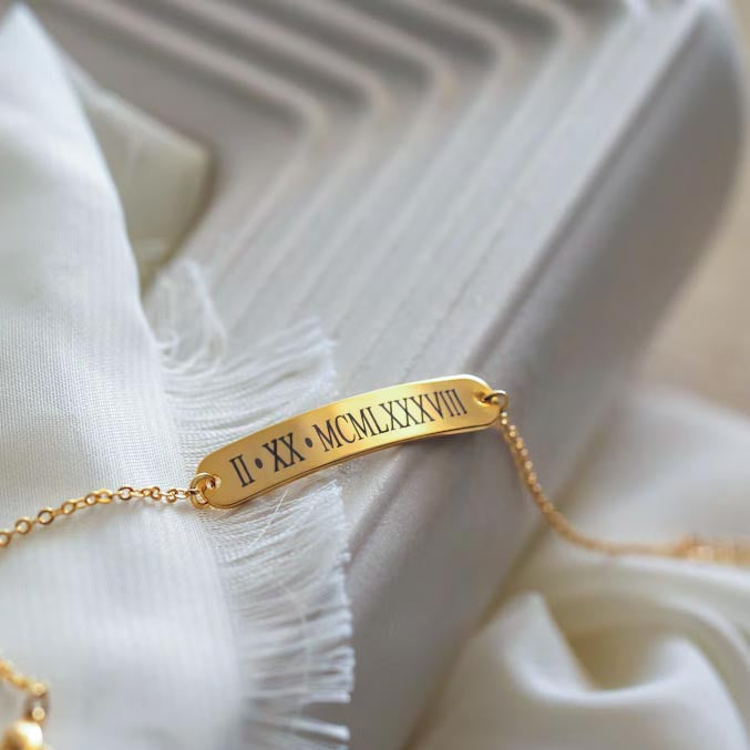 Gold-tone bar bracelet engraved with Roman numerals resting on white fabric beside a sculptural tray