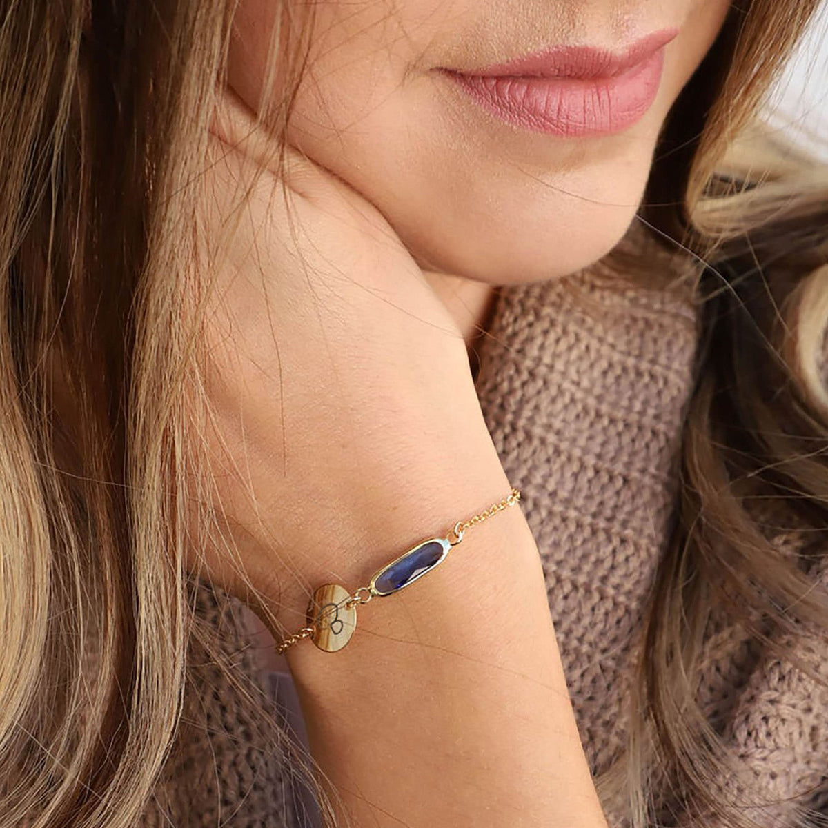 Gold-tone bracelet with engraved B initial disc and blue elongated stone worn on forearm beside a beige knit sweater