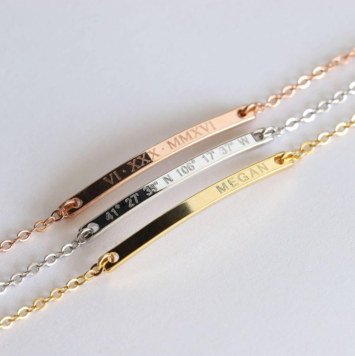 Three engraved bar bracelets in rose-gold, silver, and gold tones with Roman numerals, coordinates, and the name Megan