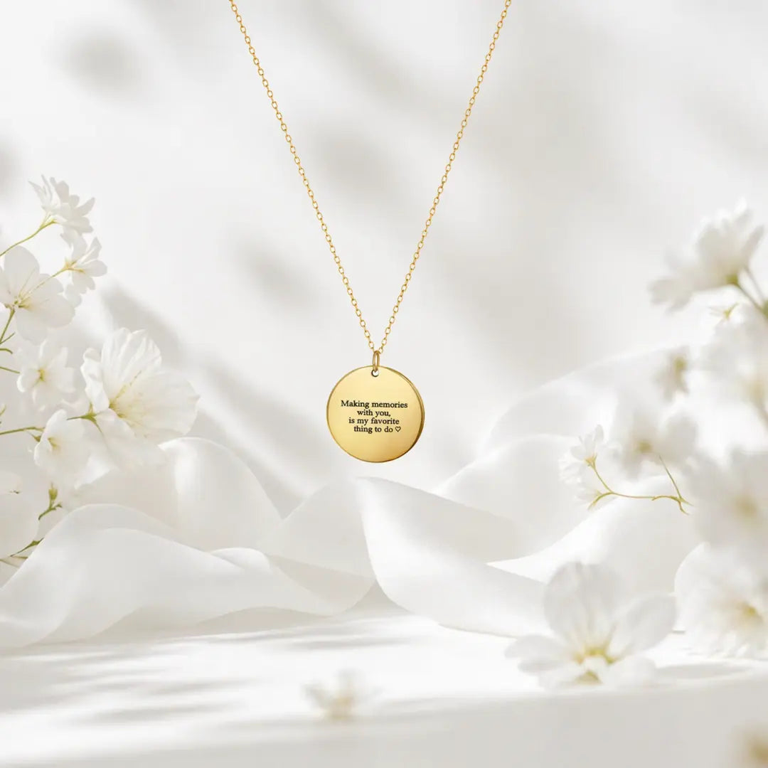 Gold-tone round message disc necklace displayed on white fabric with white flowers