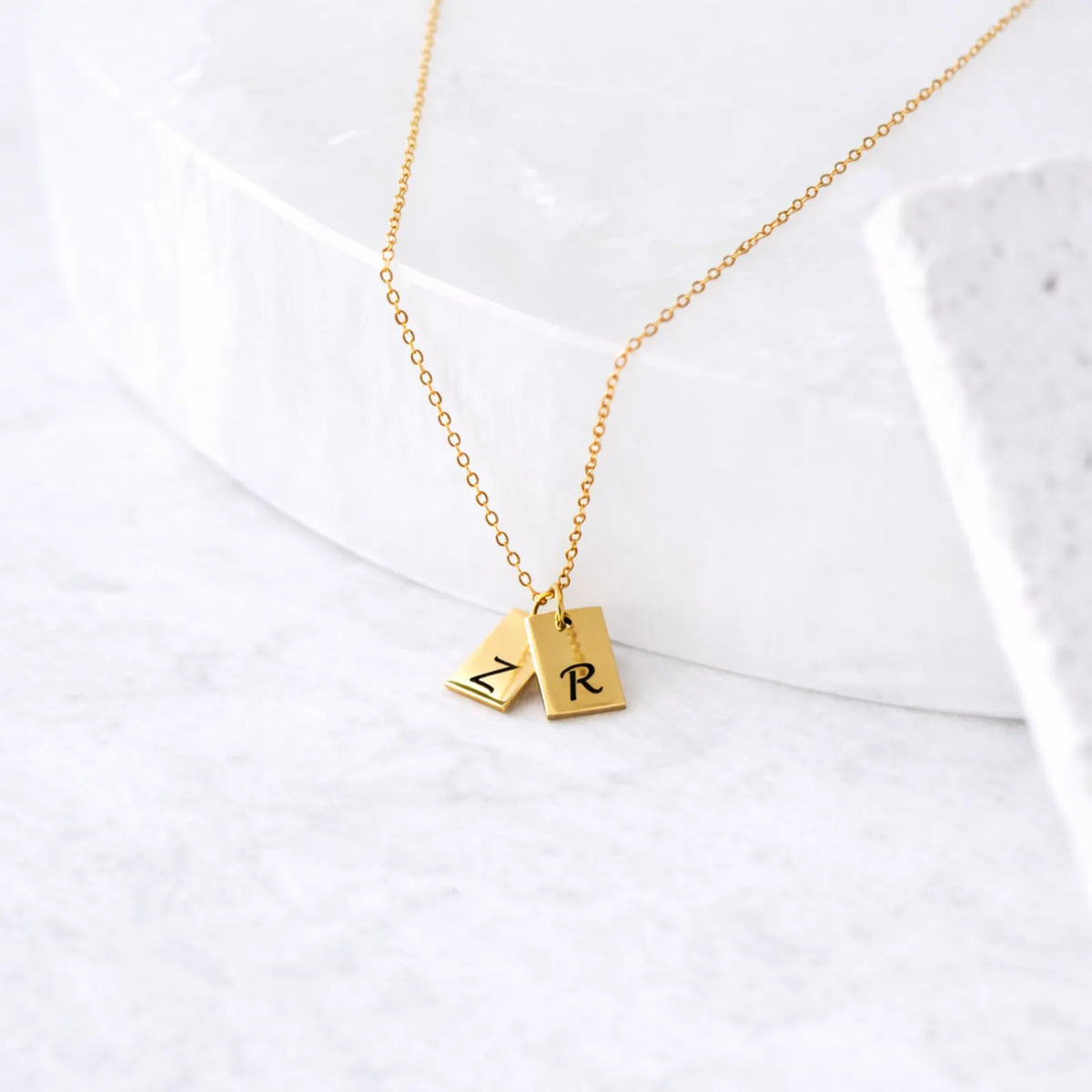 Gold-tone necklace with two rectangular tag pendants engraved N and R, displayed on a white surface