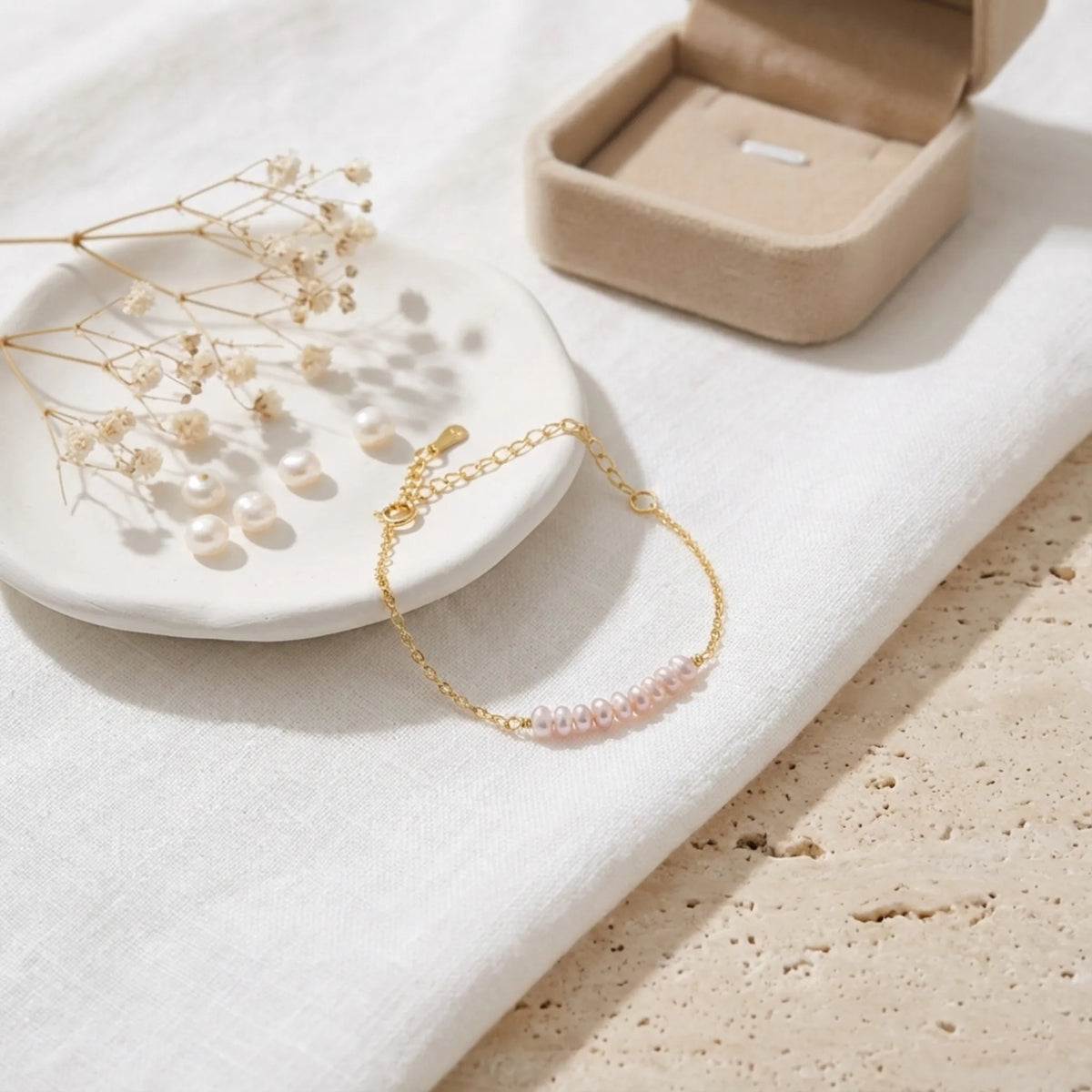 Gold-tone bracelet with pale pink oval beads beside a jewelry box and dried flowers on white fabric