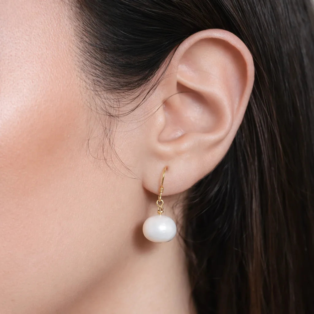 Gold-tone dangle earring with a white round drop, worn on the ear against dark hair
