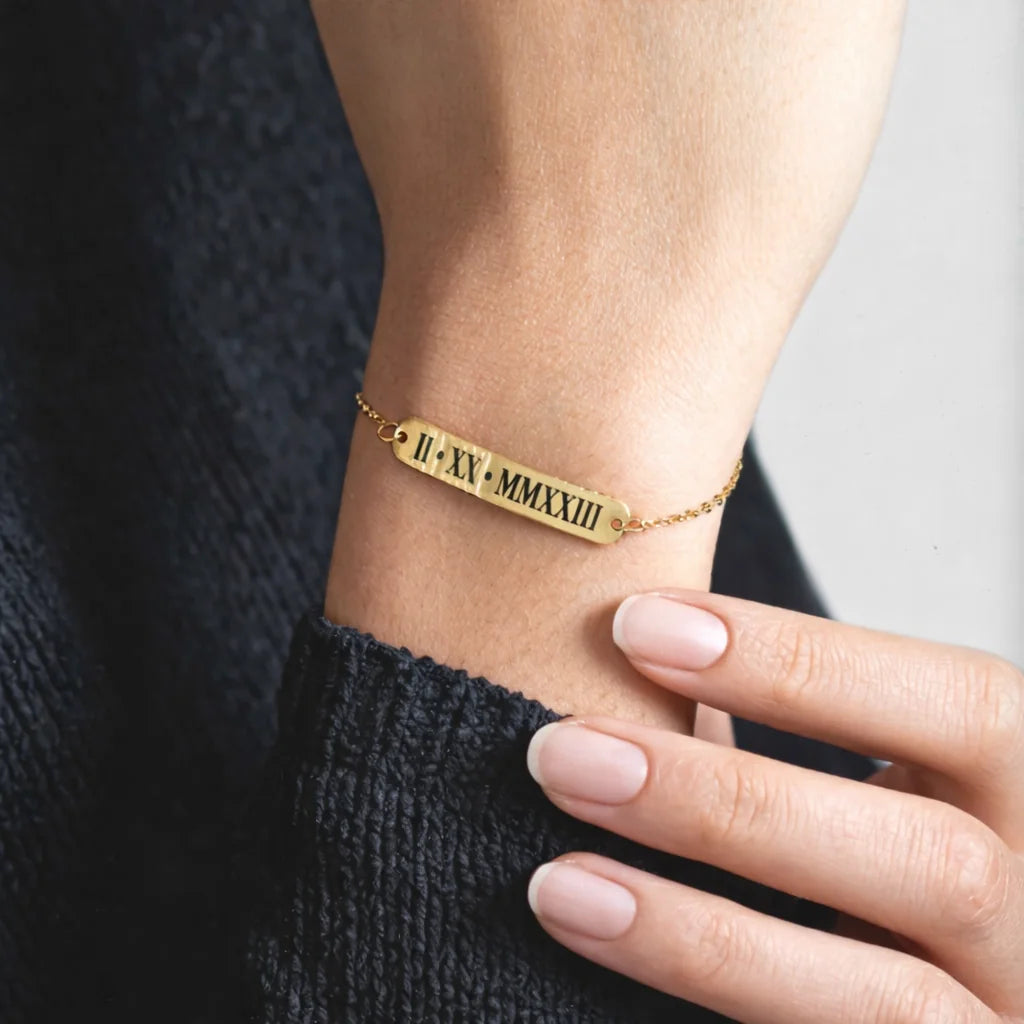 Gold-tone bar bracelet engraved with Roman numerals II XV MMXIII worn on a wrist