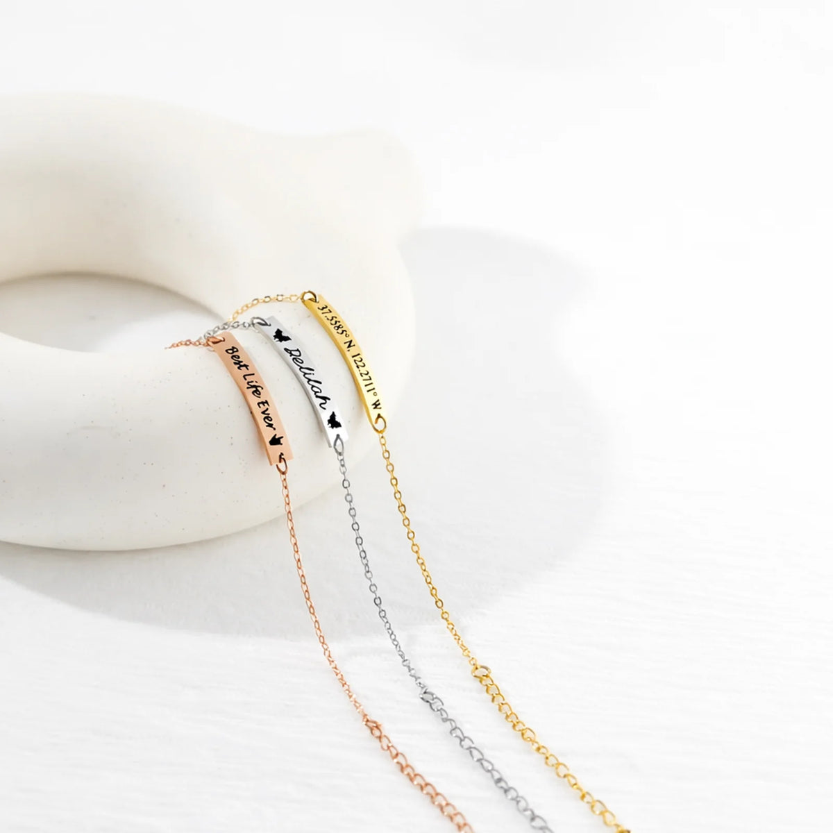 Three bar bracelets in rose-gold, silver, and gold tones with personalized engravings on a white sculptural surface