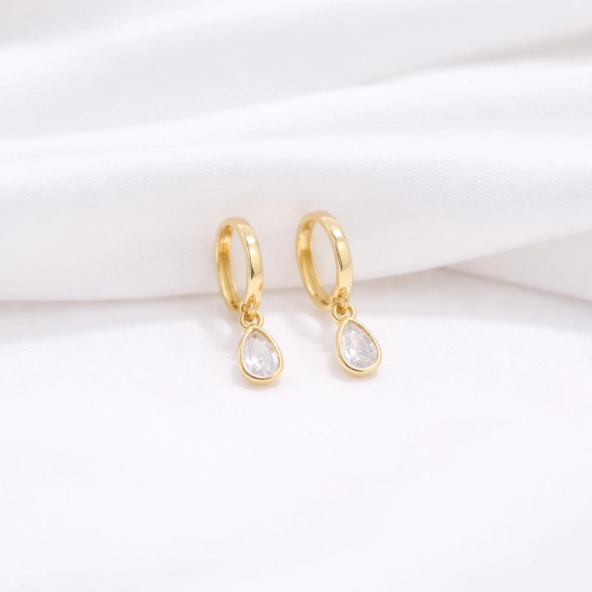 Gold-tone dangle huggie hoop earrings with clear teardrop stones displayed on softly folded white fabric
