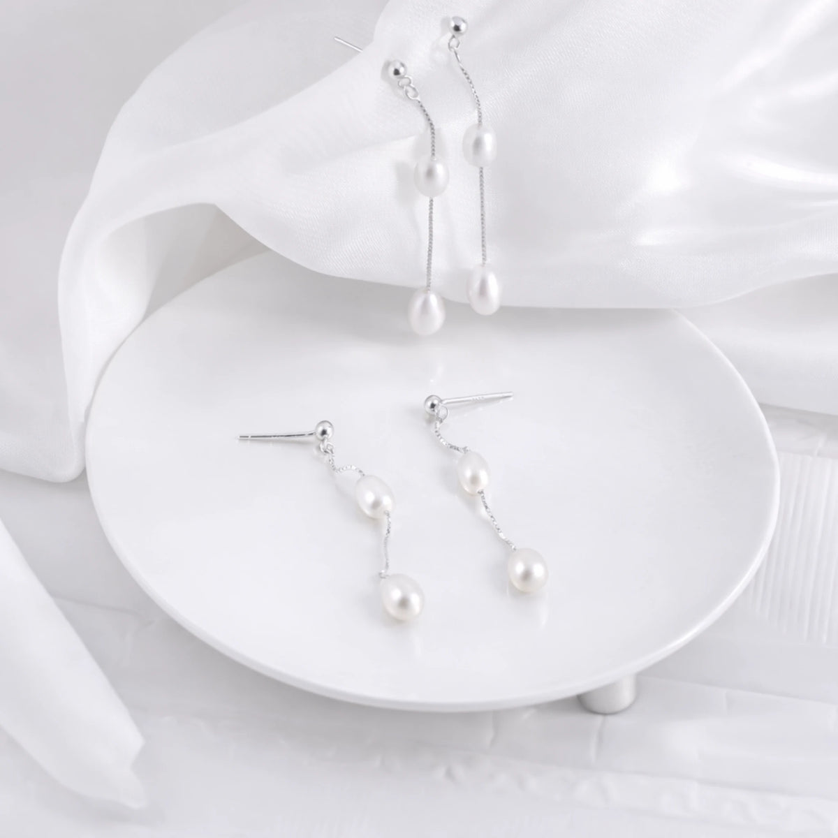 Two pairs of silver-tone drop earrings with white pearl beads arranged on a white display and satin fabric
