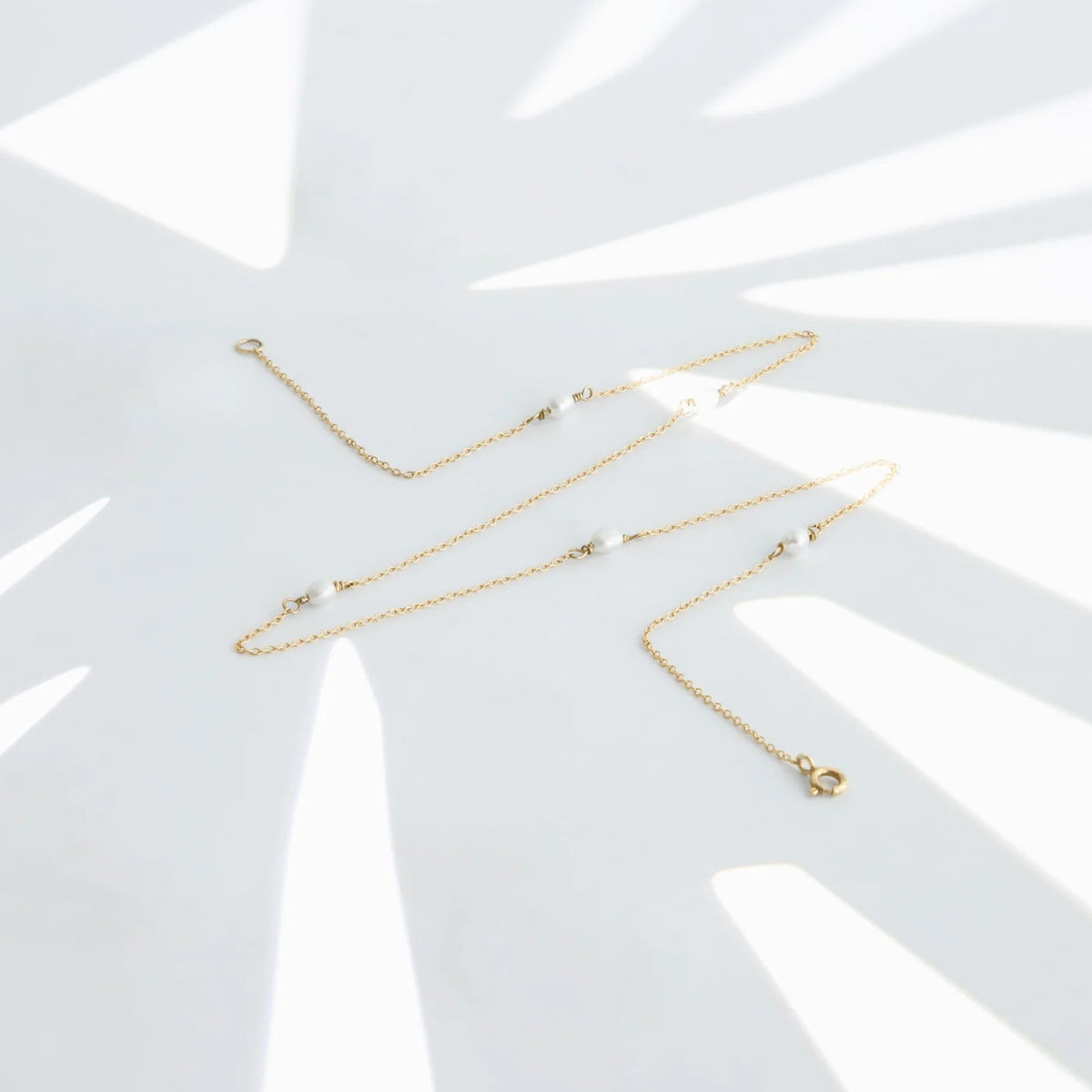Gold-tone necklace with small white oval beads arranged on a white surface with geometric shadows